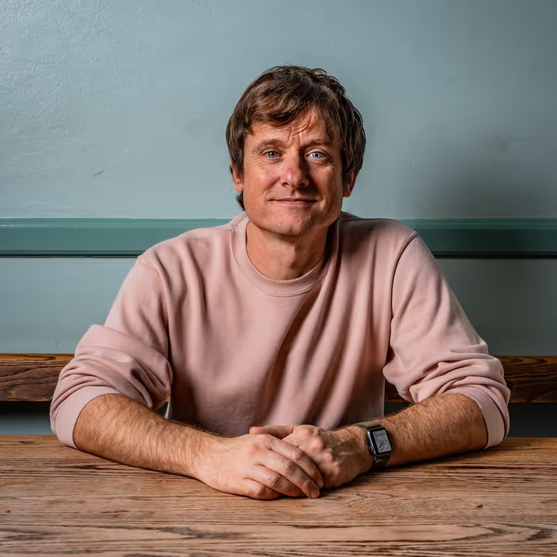 Man with short brown hair and blue eyes wearing a pink sweatshirt sitting at a wooden table with clasped hands.