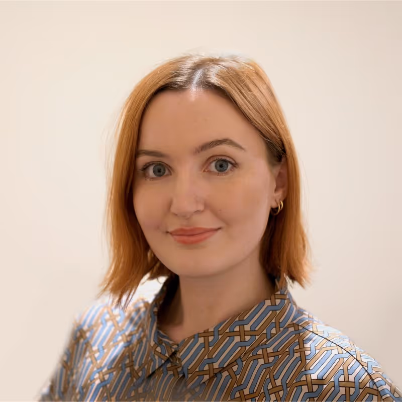 Woman with short red hair wearing a patterned shirt and small hoop earrings, smiling slightly against a plain background.