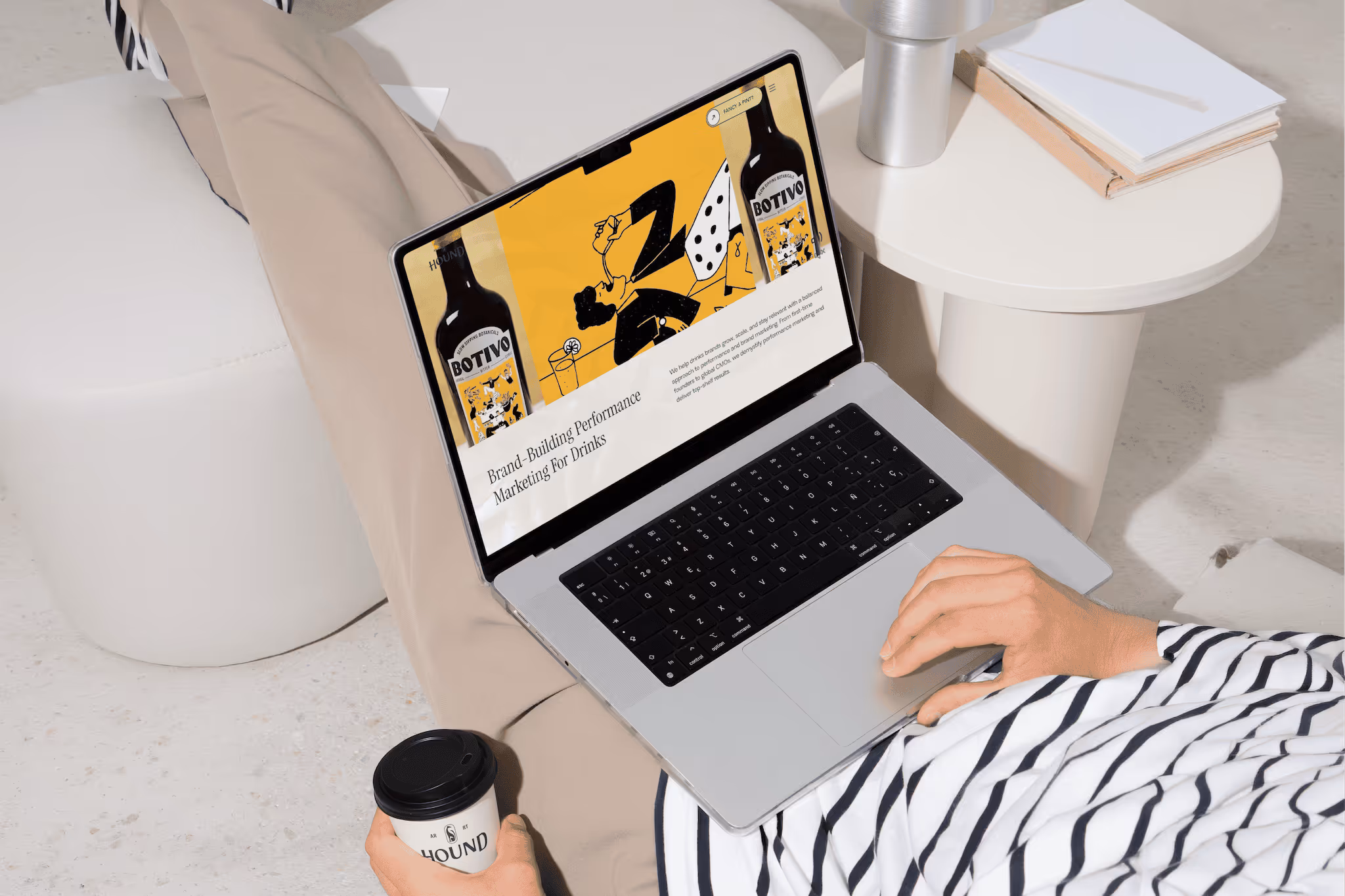 Person in striped shirt holding a coffee cup and using a laptop displaying a webpage about brand-building performance marketing for drinks.