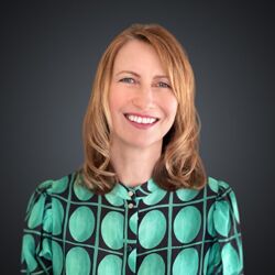 Woman with short red hair wearing a patterned shirt and small hoop earrings, smiling slightly against a plain background.