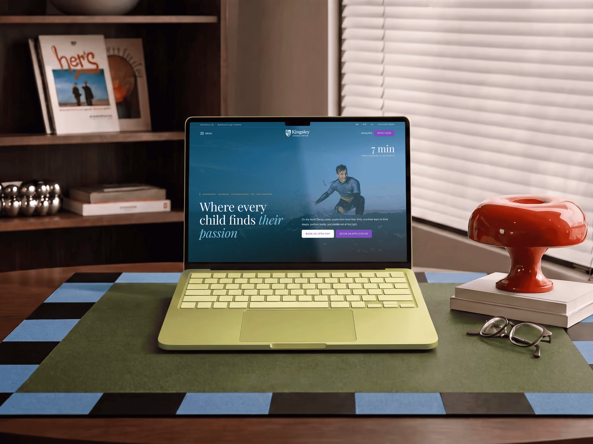 Open laptop on a table displaying a website with the text 'Where every child finds their passion' and a photo of a boy running on the screen, next to a red modern vase, a pair of glasses, and stacked books.