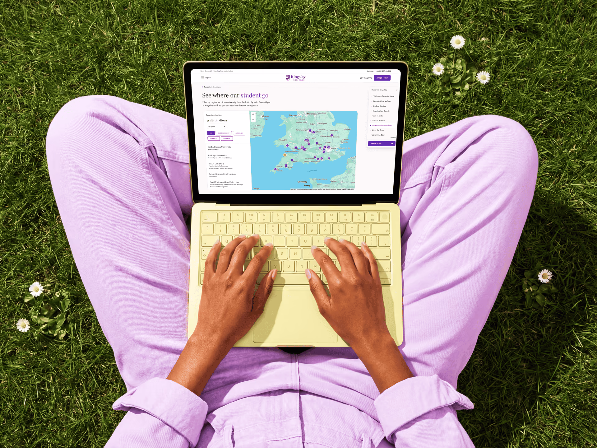 Person wearing lavender trousers sitting cross-legged on green grass with white flowers, typing on a pale yellow laptop displaying a UK map labeled 'See where our student go' with purple markers.