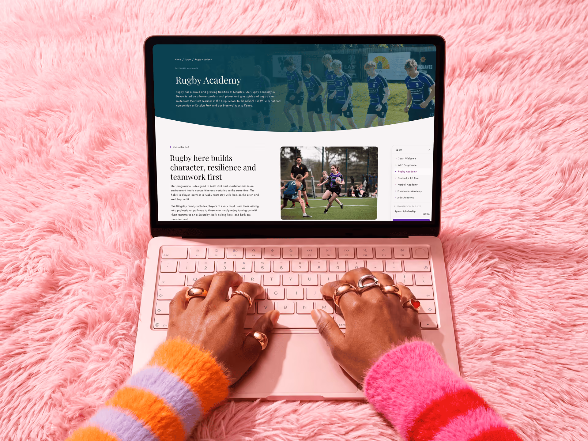Person with multiple rings on fingers typing on a pink laptop with a Rugby Academy webpage open, placed on a fluffy pink surface.