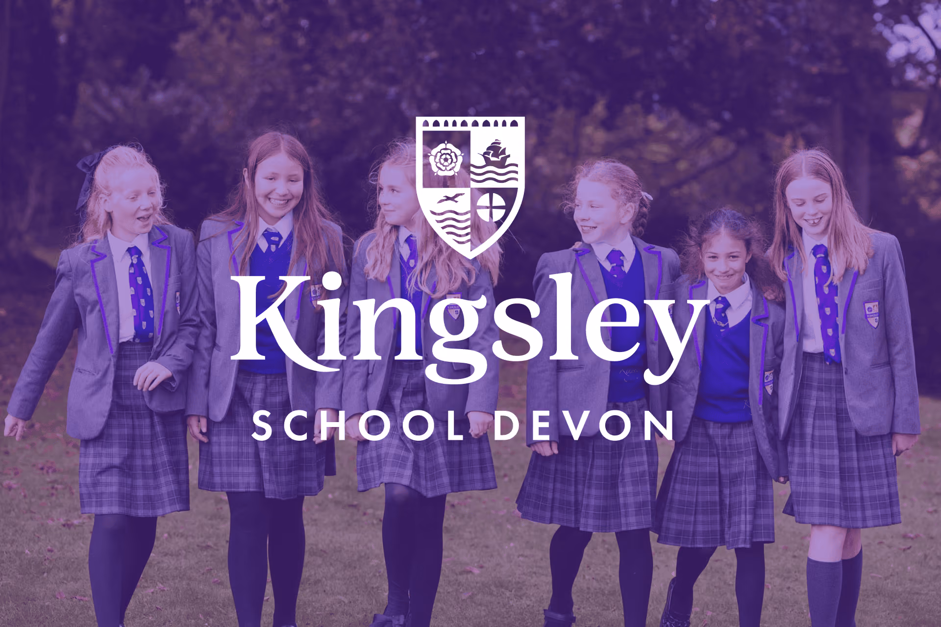 Six girls in school uniforms with grey blazers, blue jumpers, plaid skirts, and blue ties walking together outside on grass, smiling and talking.