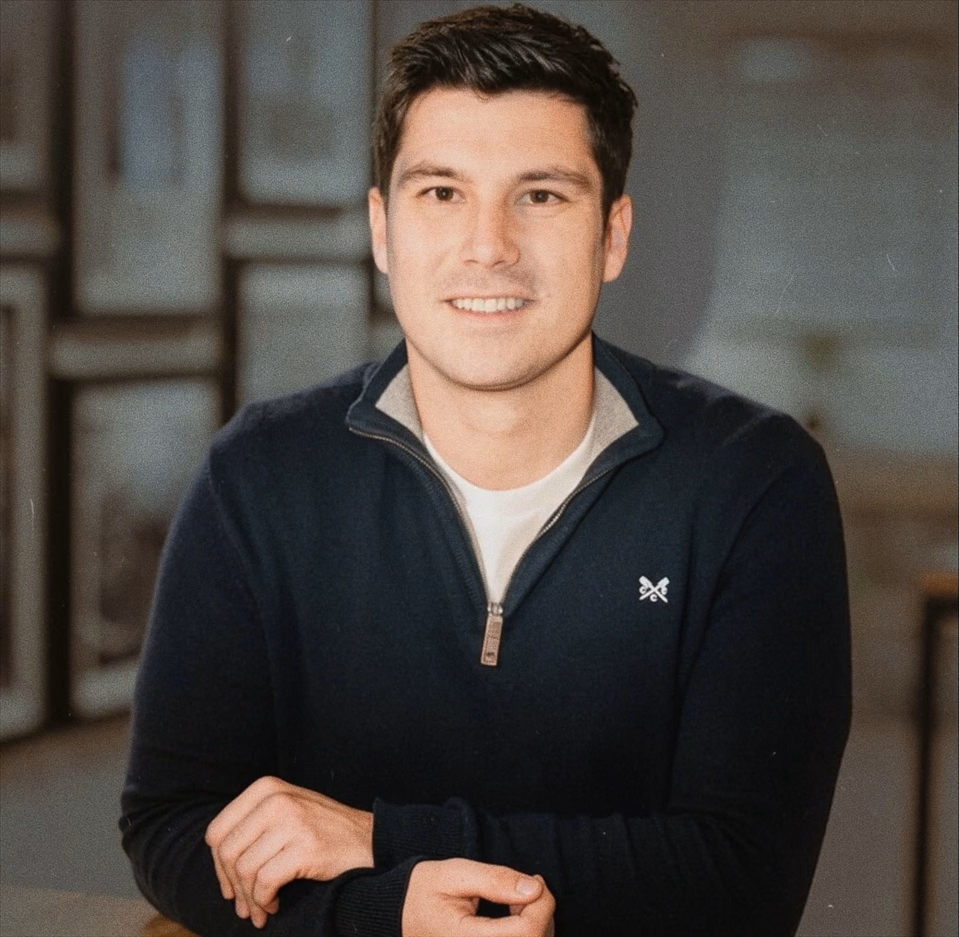 Smiling young man with short dark hair wearing a navy blue zip-up sweater over a white t-shirt, seated indoors with hands folded on a table.