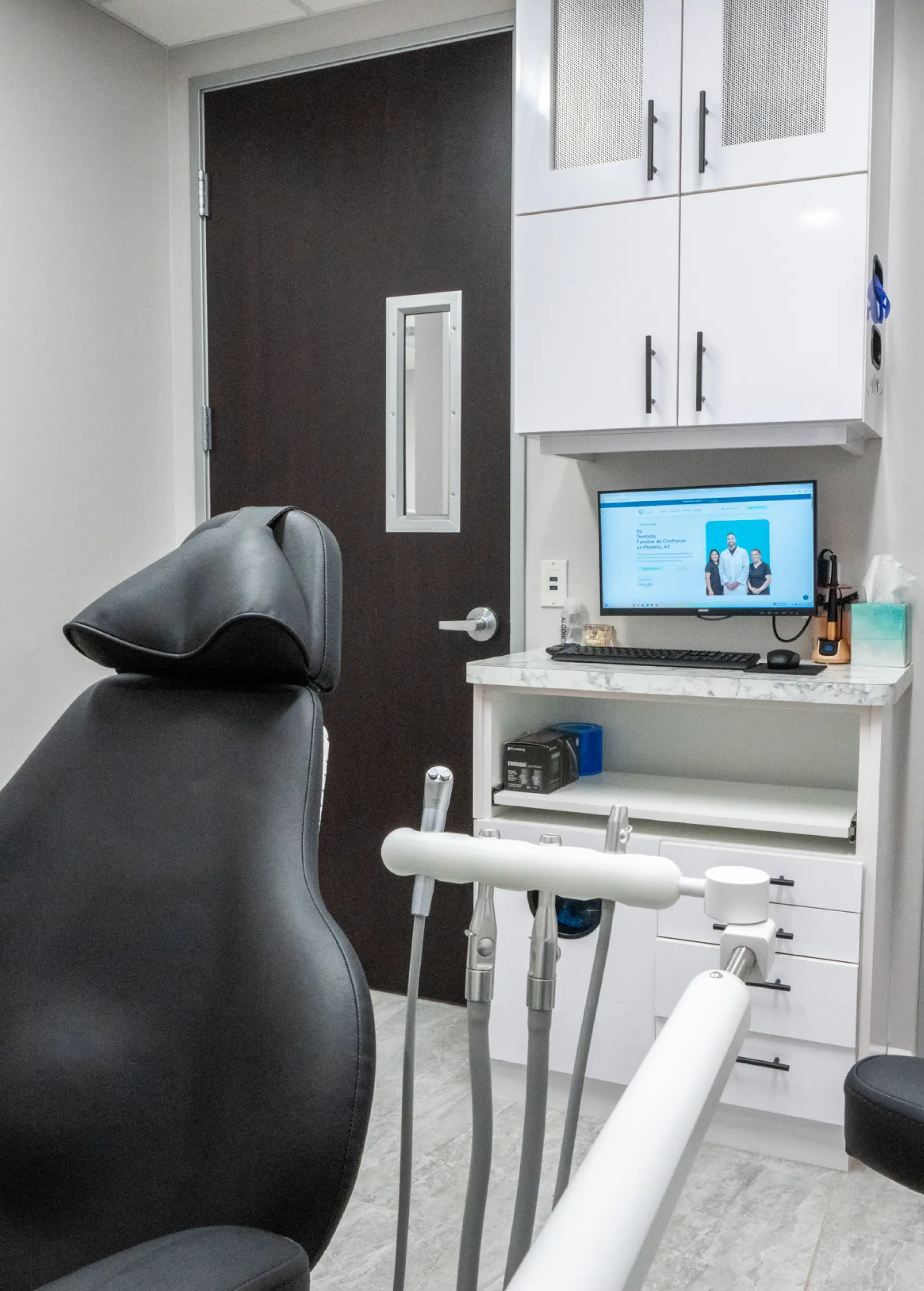 Dental exam room with a black chair, dental tools, and a computer on a white cabinet.