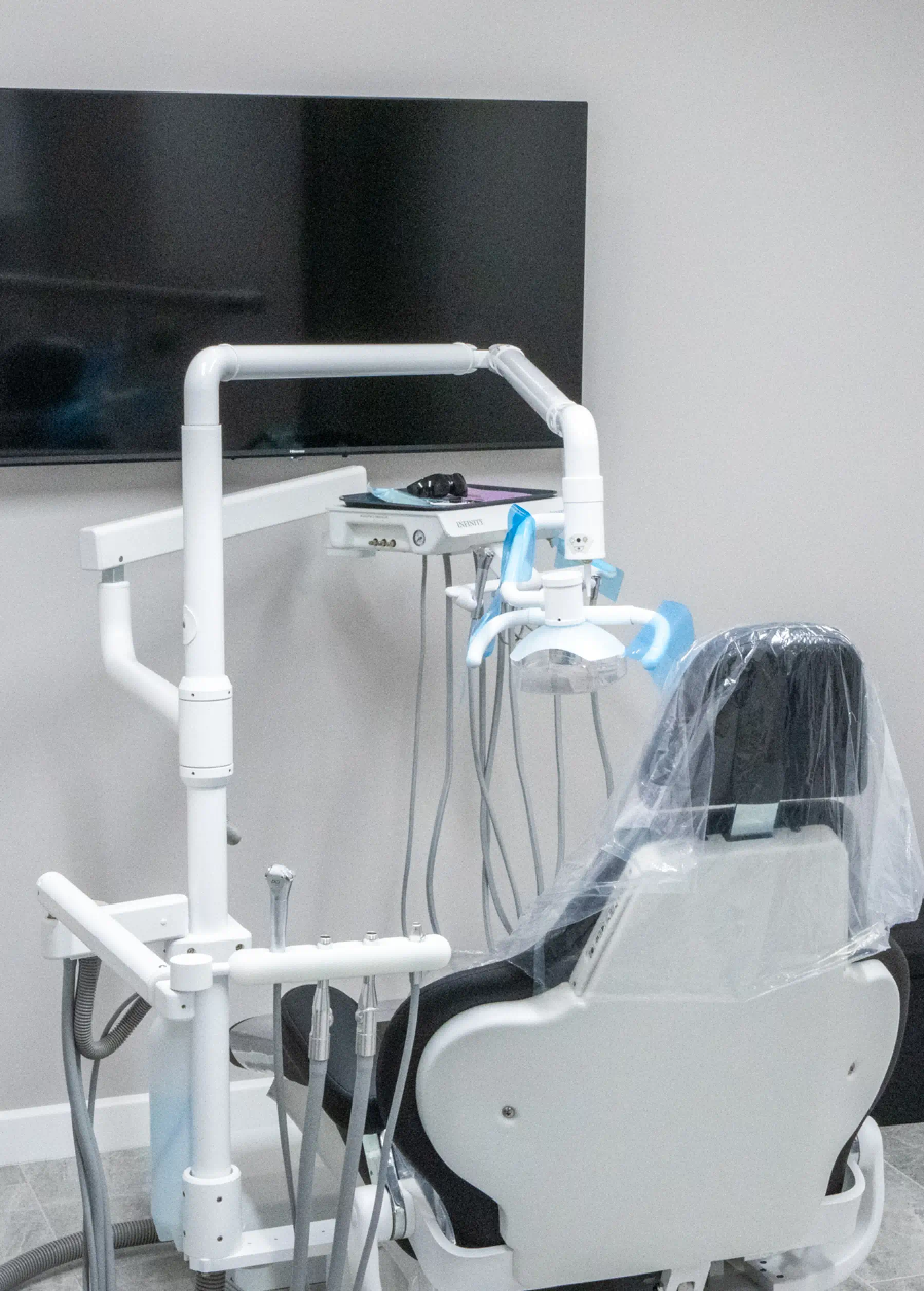 Dental chair with overhead light and instruments, covered in protective plastic, facing a television on the wall.