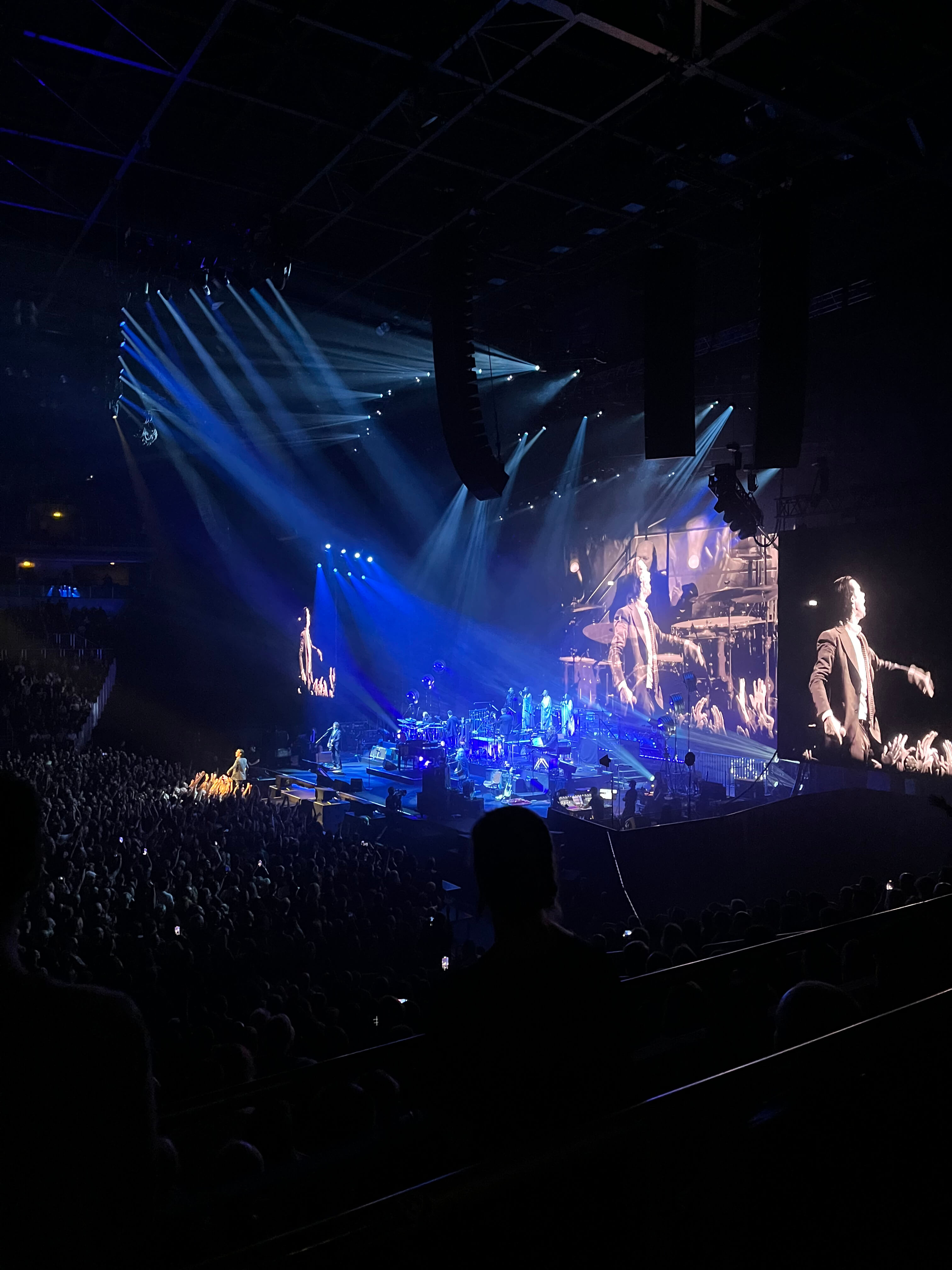 Large indoor concert with crowd, stage musicians, and a performer in a suit projected on big screens under blue stage lights.