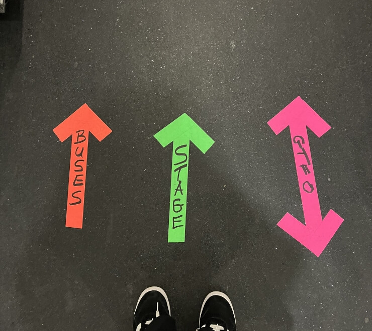 Close-up of brightly coloured fluorescent labeled arrows on the floor, pointing to 'Buses', 'Stage', and way out for technical crew direction.