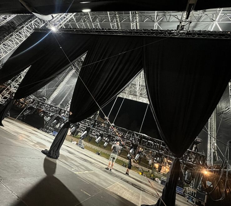 Low-angle shot from inside a large outdoor stage structure, showing trussing, moving head lights, and large black stage drapes being adjusted by two technical crew members during pre-production.