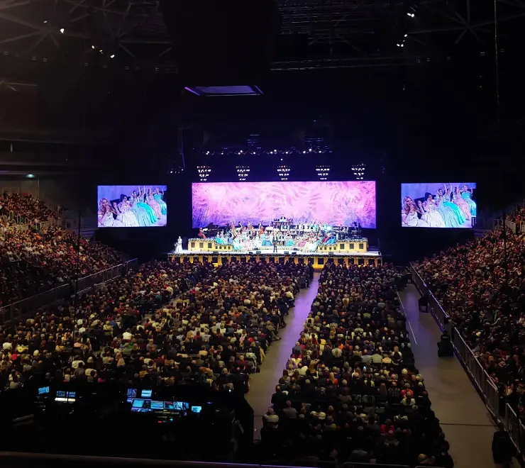 Large audience seated in a concert hall watching a live orchestra performance with a colorful backdrop and two large screens showing dancers in elaborate dresses.