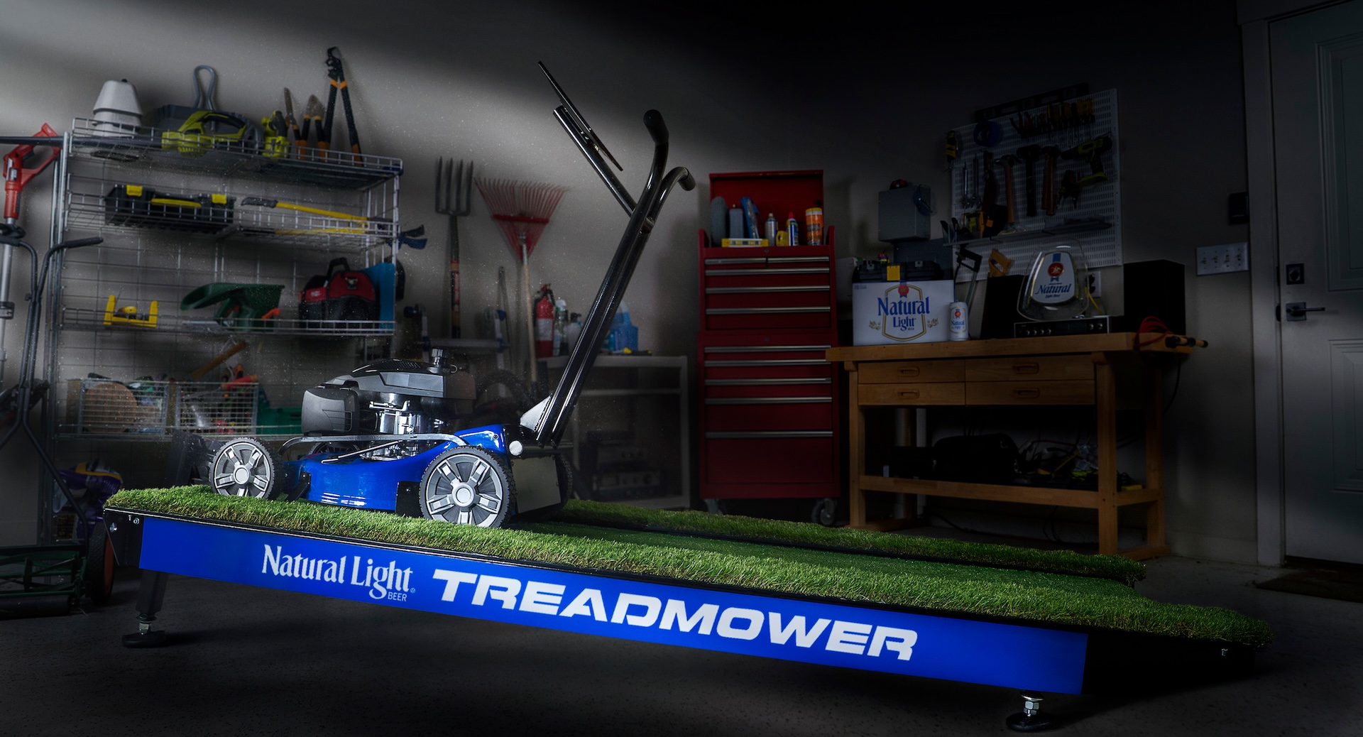The Natural Light TreadMower, featuring a blue lawnmower positioned on a treadmill track with a grass-covered surface inside a home garage workshop.