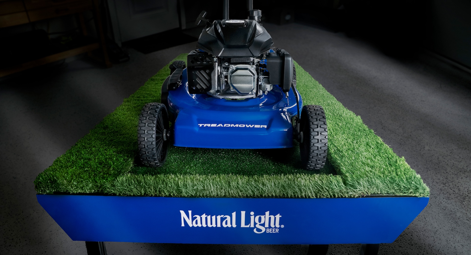 A front view of the Natural Light TreadMower, showing the blue lawnmower deck and wheels on a vibrant green, grass-covered treadmill track.