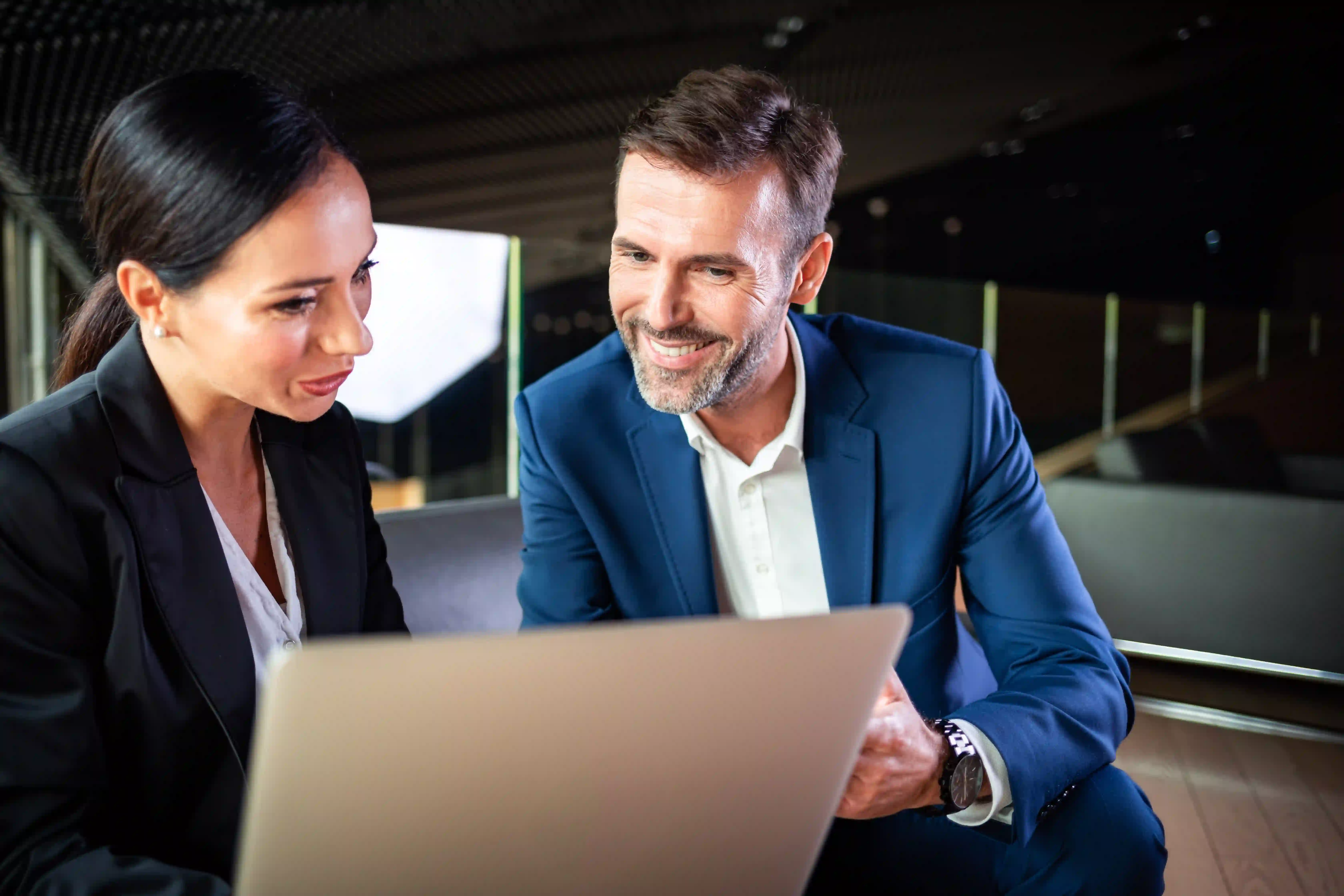 A man and woman in business attire are focused on a laptop, collaborating on a project in a professional setting.