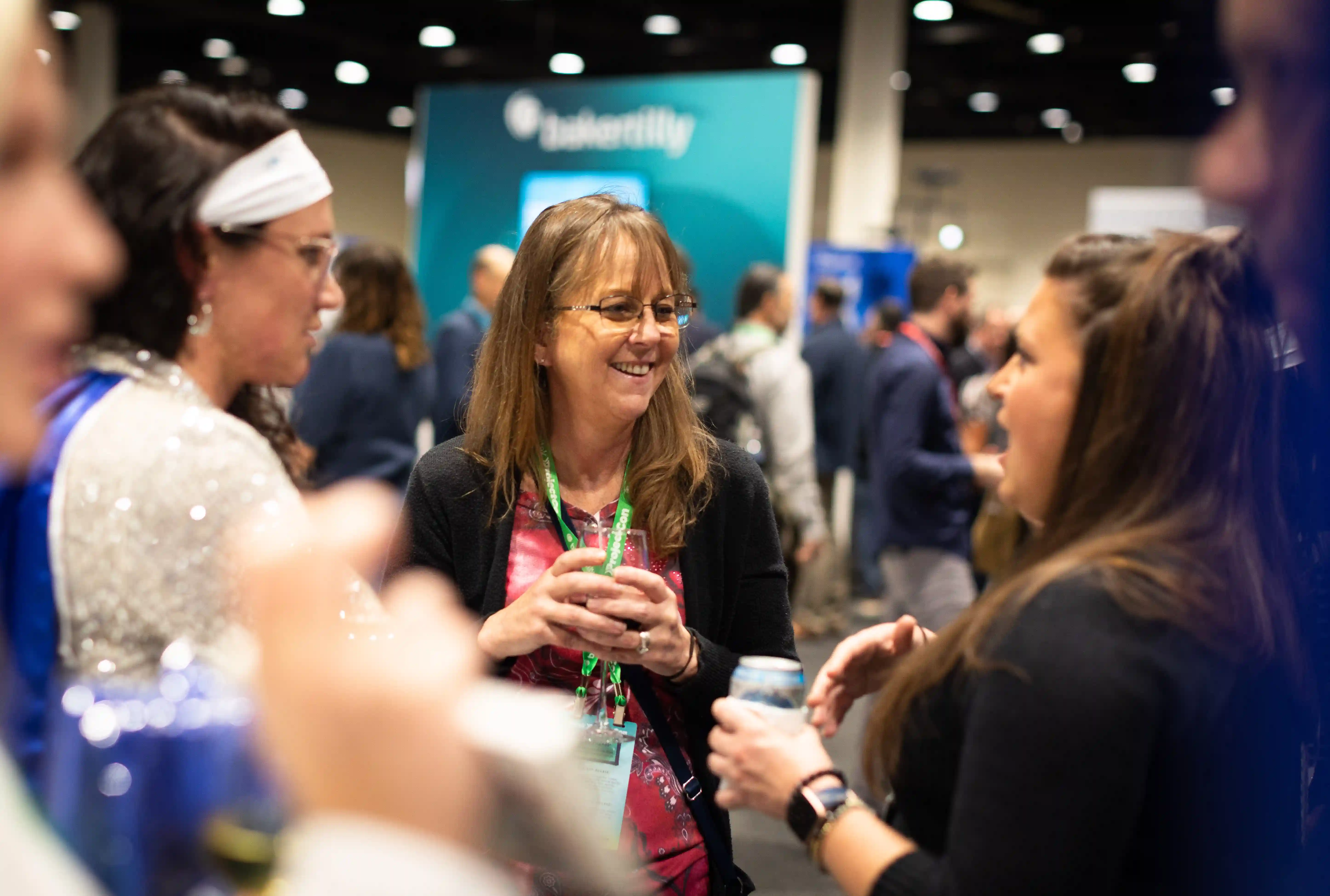 Two women engaged in conversation at a convention, surrounded by attendees and displays, showcasing a vibrant atmosphere.