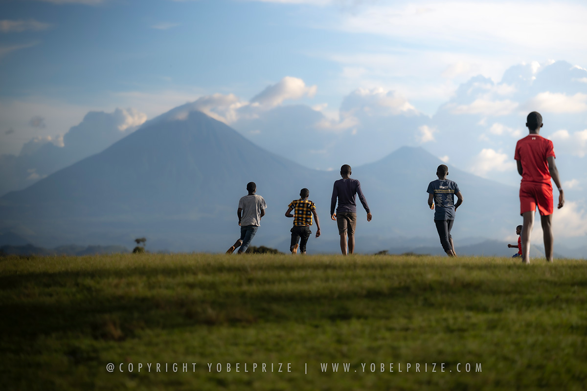 Young Ugandan boys playing soccer, running energetically across an open field with dramatic volcano peaks in the background.