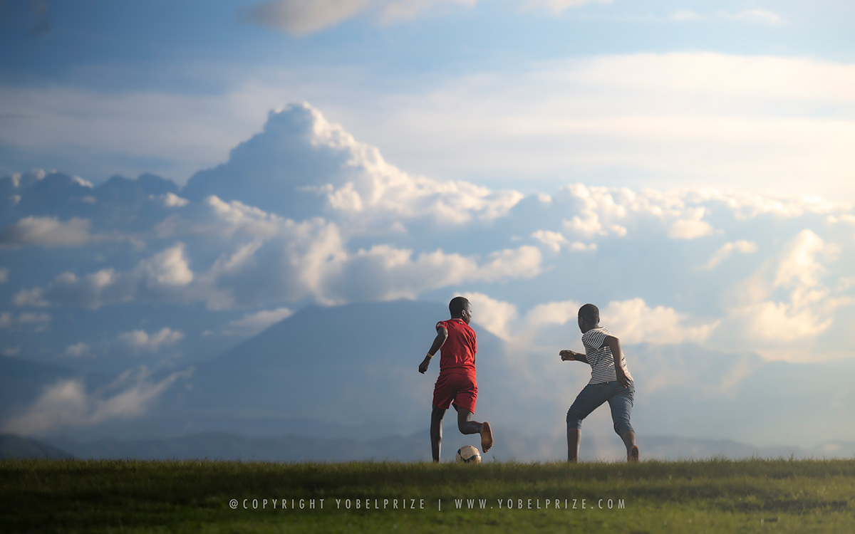 Two young boys energetically playing soccer on an open grassy field, silhouetted against a backdrop of expansive clouds and distant mountainous landscapes in Uganda.
