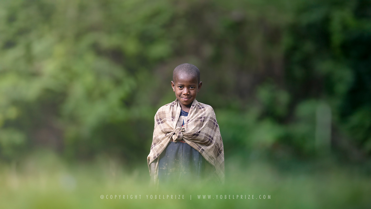 Boy Smiling in Shawl: A young Ugandan boy wrapped warmly in a beige shawl stands in a lush green landscape, smiling gently towards the camera.