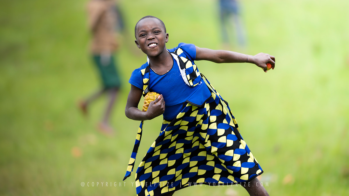 Joyful Girl Playing: An exuberant Ugandan girl wearing a vibrant blue and yellow patterned dress energetically plays outdoors, radiating pure joy.