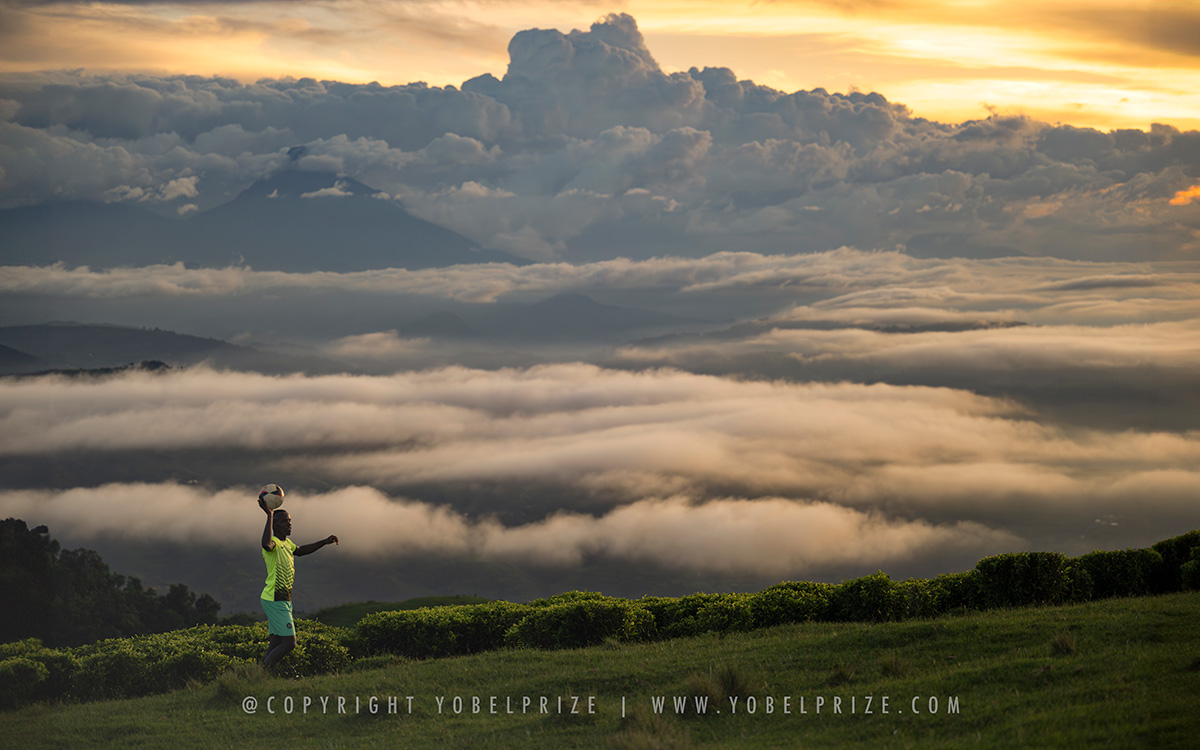 Boy Playing Soccer Above Clouds: A young Ugandan boy skillfully balances a soccer ball against an ethereal backdrop of rolling clouds and mist-covered mountains at sunset.