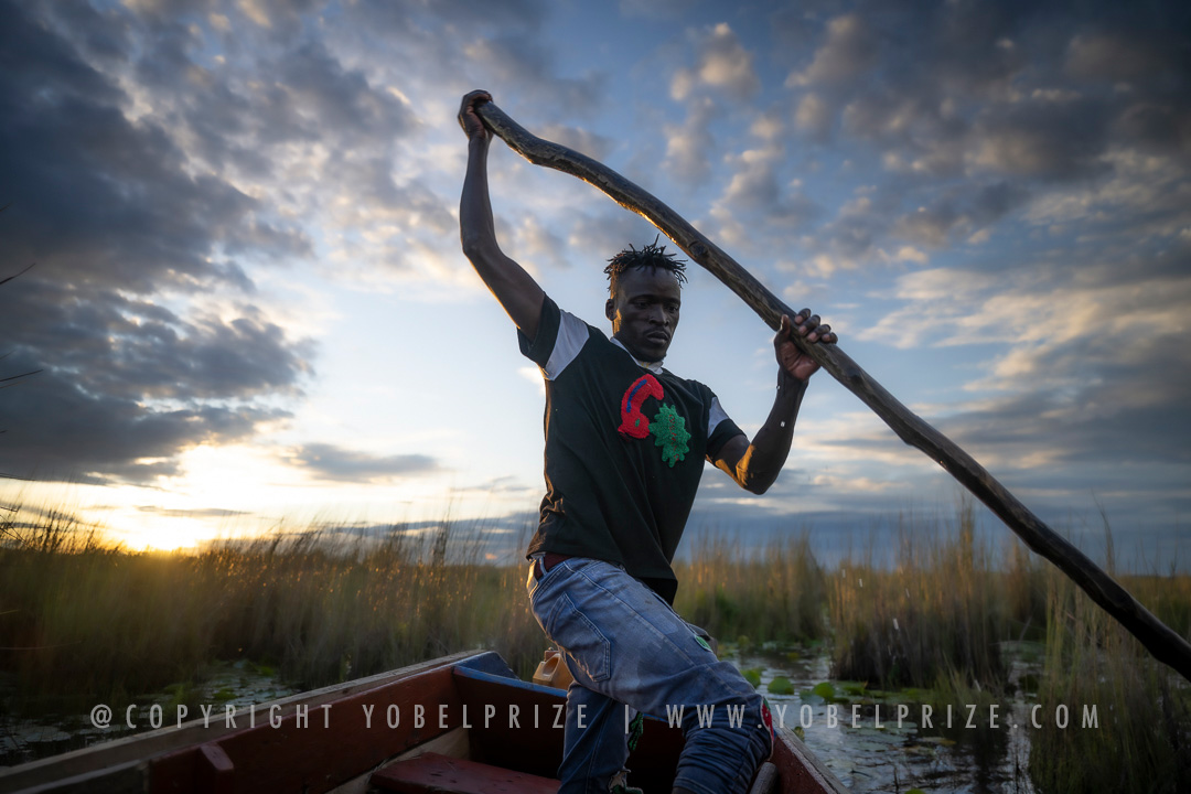 A local Ugandan man skillfully steering a canoe using a long wooden pole, captured dramatically against a vivid sunset sky with reflections shimmering on the marshy water of Mabamba Swamp.
