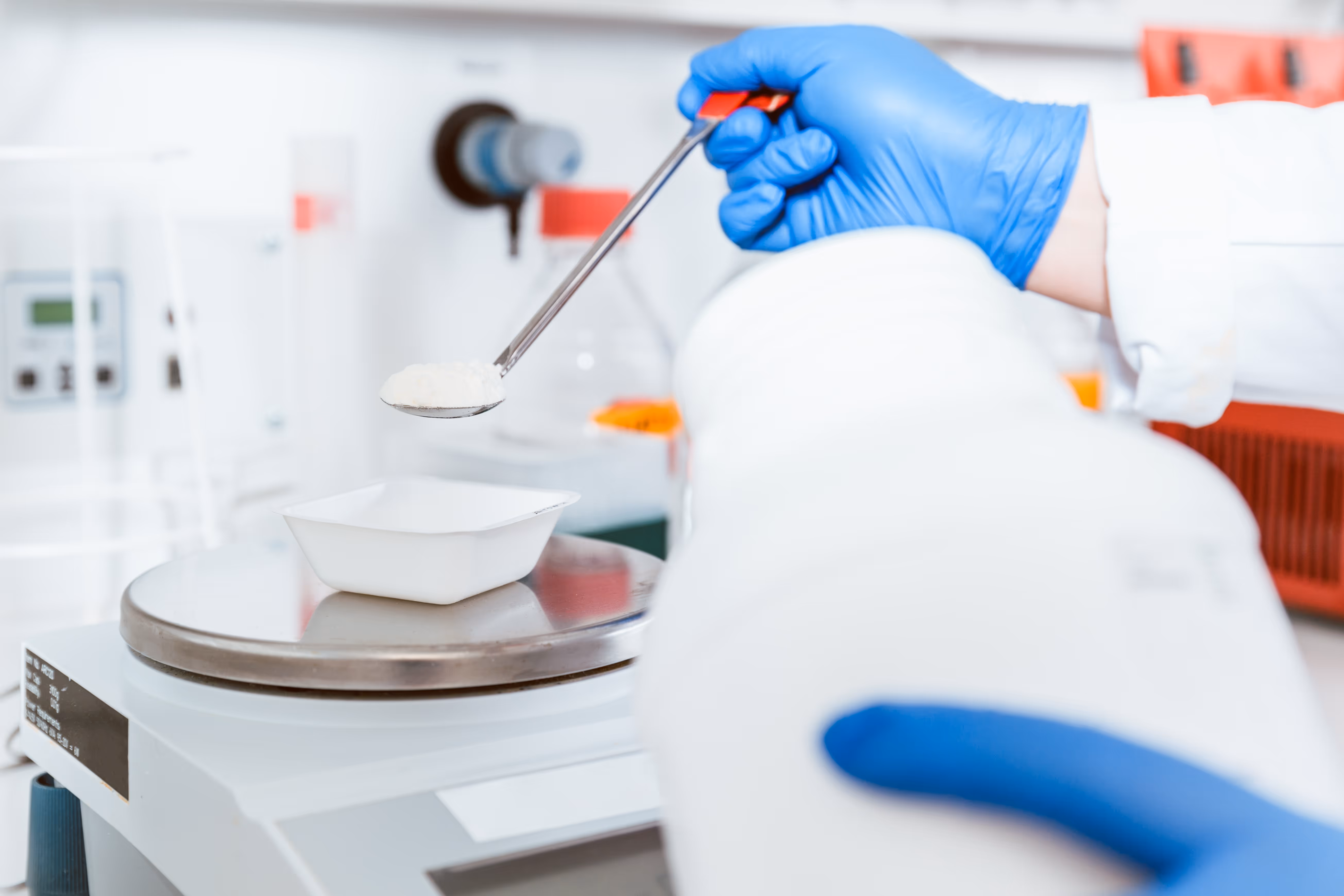 Lab technician wearing blue gloves measuring white powder on a spoon over a scale in a laboratory.