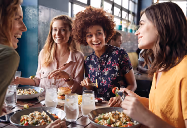 Four women smiling and enjoying a meal together at a restaurant table with plates of food and drinks.