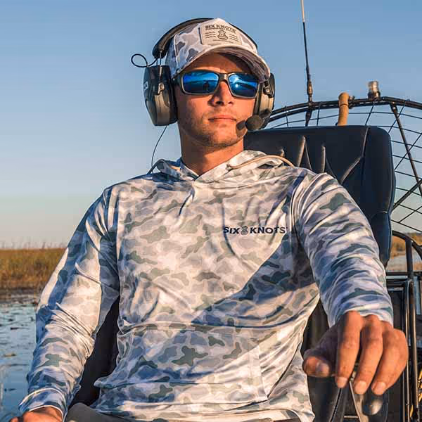 Man airboating in sunglasses, wearing the Six Knots water-resistant Coastal Camo Snapback Hat and Wetlands-pattern Chesapeake Performance Hoodie — lightweight, moisture-wicking boating apparel