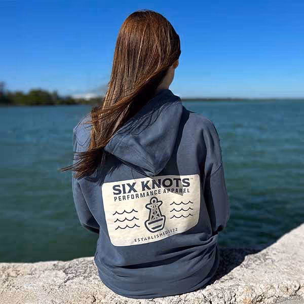 Rear view of woman sitting by the water wearing the Six Knots Making Waves Classic Hoodie in navy. This ultra-soft unisex hoodie is crafted from a breathable, premium cotton-blend fabric and features a relaxed fit for everyday comfort. Ideal for layering on cool coastal mornings, casual weekends, travel days, and laid-back seaside adventures.