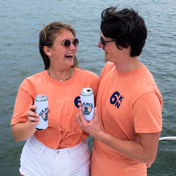 A man and a woman smiling at one another near the water while holding canned beverages and wearing Six Knots Weekender Classic T-Shirt in Terracotta color.