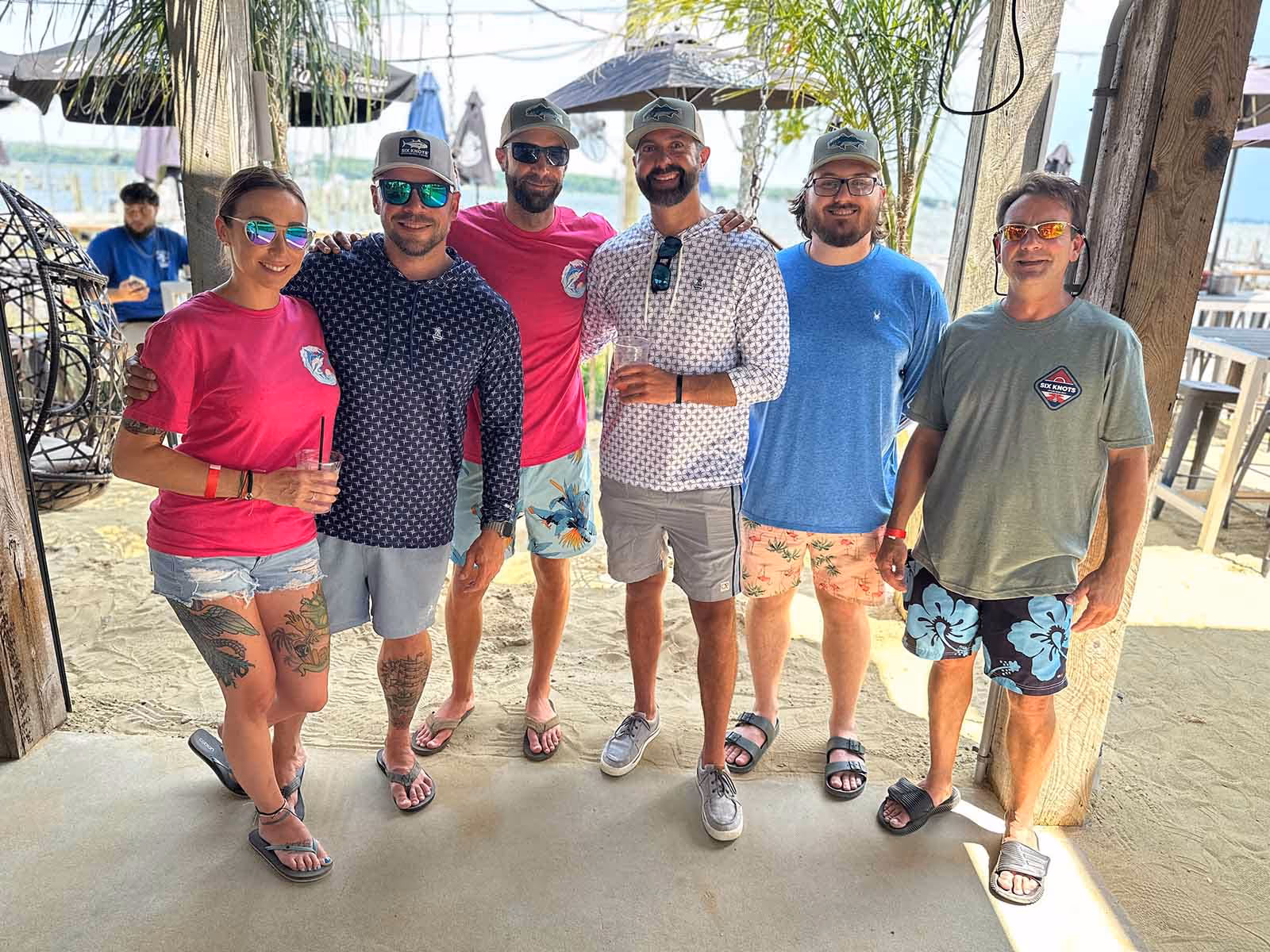 Group of friends at a dockside bar wearing Six Knots coastal apparel, smiling and holding drinks—showcasing casual boating lifestyle, social waterfront gatherings, and nautical-inspired clothing.