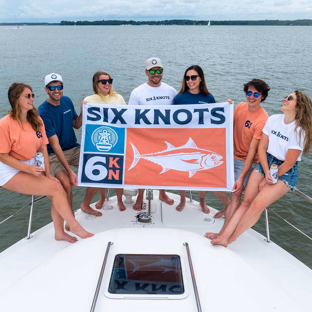 A group of men and women smiling on the bow of a powerboat holding up a Six Knots Tuna Troller flag featuring multiple logos and a tuna fish graphic inside an orange box. Showcasing boating lifestyle, coastal apparel branding, and offshore fishing culture.