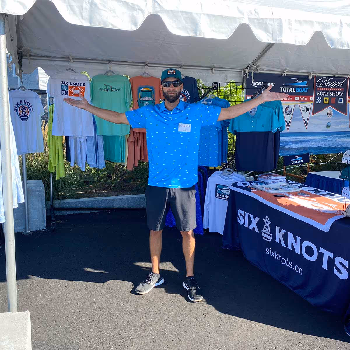 Man standing in the Six Knots booth at the Newport International Boat Show, arms outstretched proudly displaying the latest coastal apparel and performance gear.