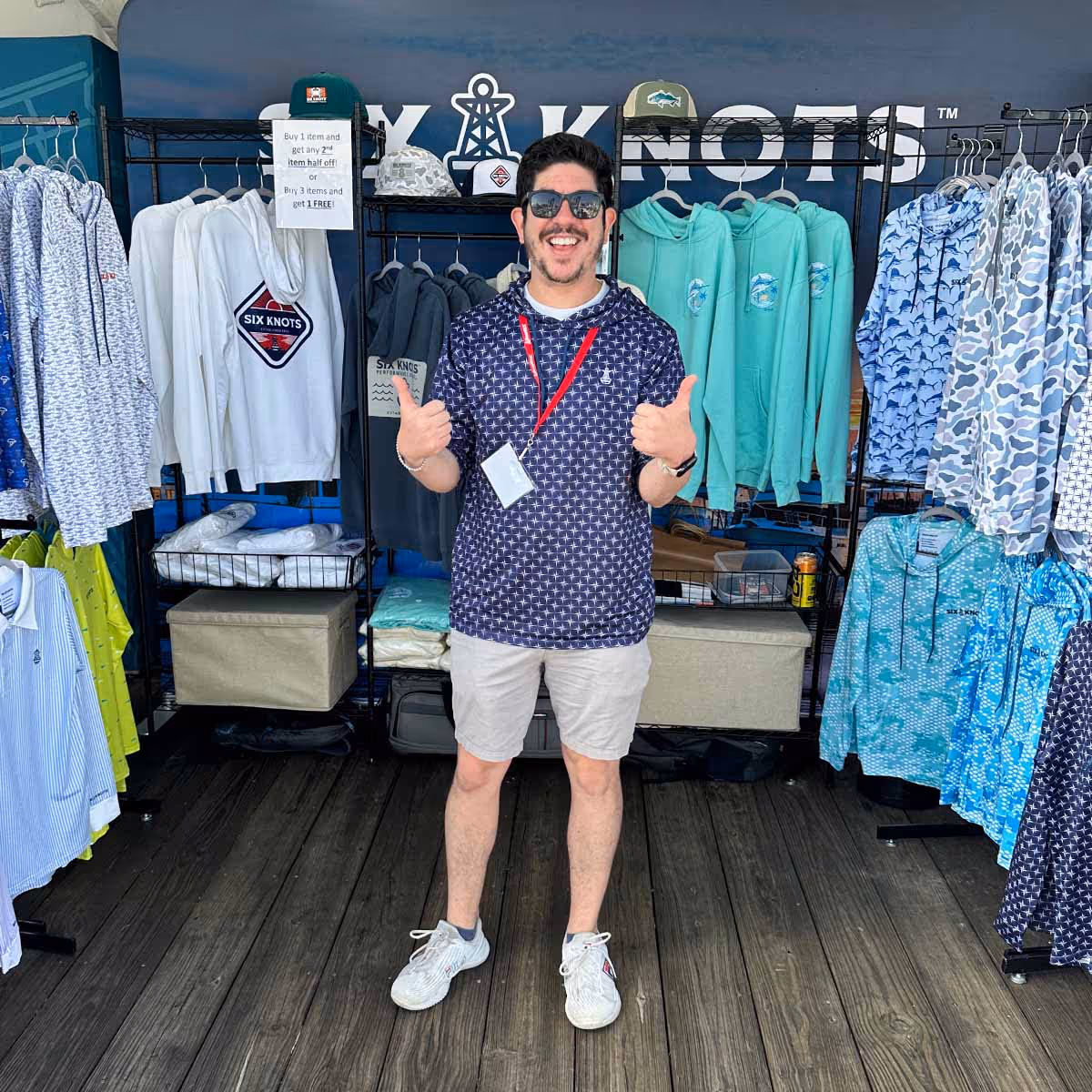 Man standing in the Six Knots event booth giving a thumbs up, wearing the Navy True North Performance Hoodie and showcasing the latest apparel collection.