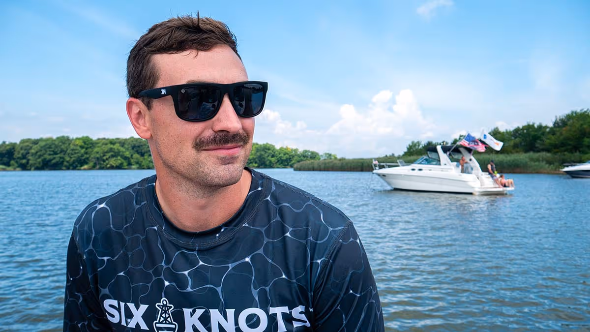 Man sitting on a powerboat wearing sunglasses and a black Six Knots Deep Water Men's Performance Crew. This lightweight, moisture-wicking fishing shirt features a dynamic water-inspired pattern and provides UPF sun protection. Designed for saltwater boating, coastal fishing, and active days on the water, it delivers breathable comfort and all-day performance under the sun.