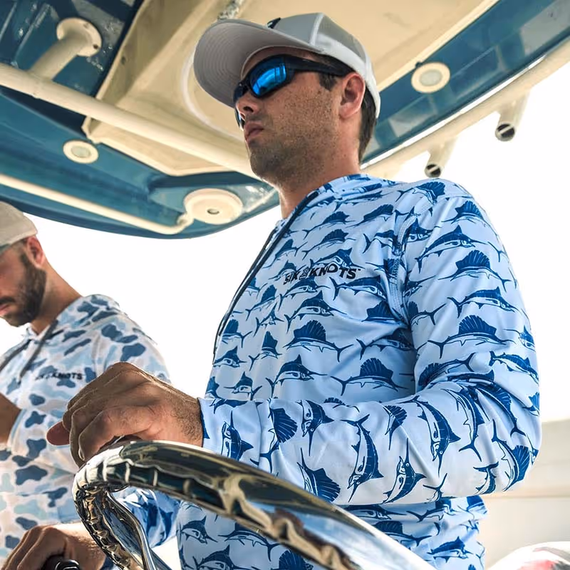 Man piloting a powerboat while wearing the Six Knots Swords and Sails Performance Hoodie — lightweight, UV-protective boating apparel