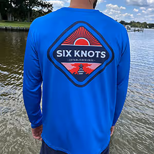 Rear view of man standing near the water wearing the Six Knots Red Sky At Night Men’s Performance Crew in vibrant royal blue, featuring a bold red and navy ‘Six Knots Established 2022’ back badge — lightweight, breathable, and moisture-wicking fishing shirt designed with UPF sun protection and quick-dry fabric; perfect for saltwater fishing, offshore boating, and all-weather coastal adventures.