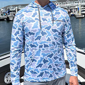 Man on a boat in a marina wearing the Six Knots Coastal Camo Performance Hoodie in Chesapeake colorway. This lightweight, moisture-wicking men's hoodie features breathable, quick-dry fabric and a bold blue and gray coastal camouflage pattern. Designed for all-day comfort and sun protection, it's perfect for boating, saltwater fishing, outdoor adventures, and year-round coastal lifestyle wear.
