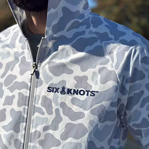 Close-up front view of Six Knots Coastal Camo Rain Jacket showing embroidered logo and waterproof zipper — lightweight, water-resistant outerwear designed for boaters, fishermen, and coastal adventurers