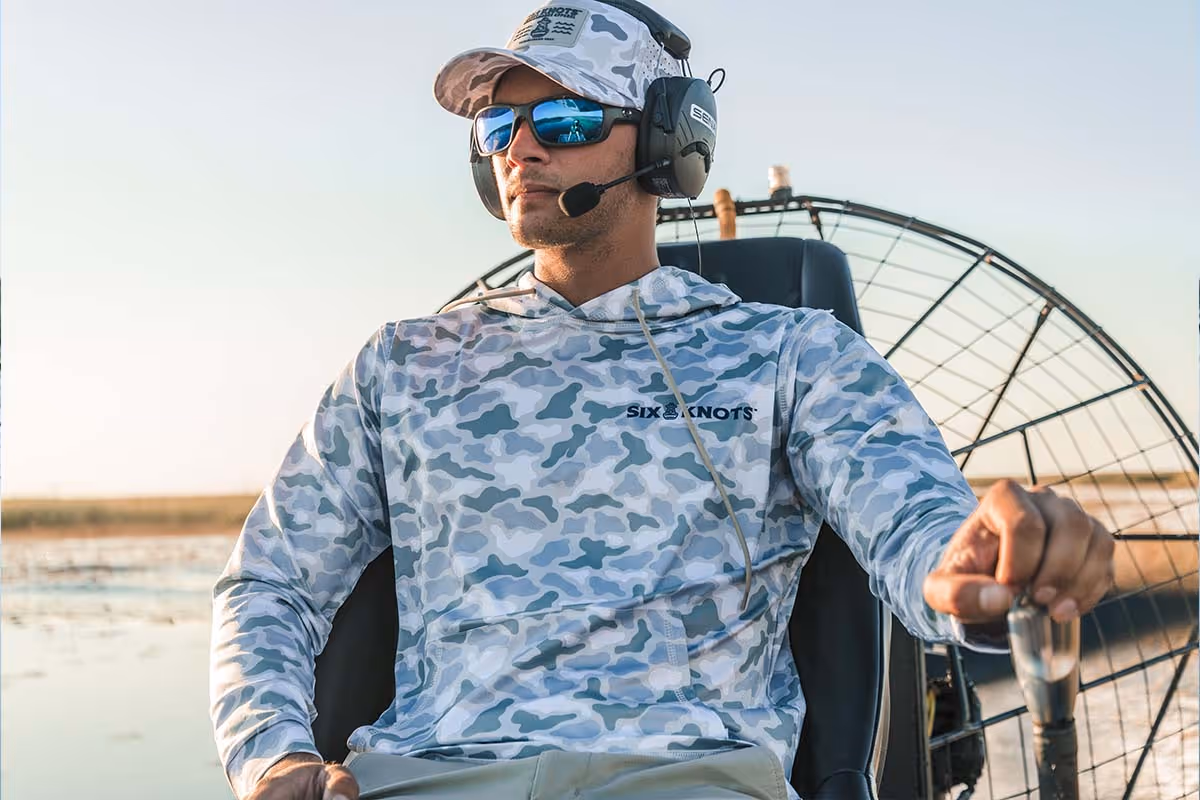 Man airboating in sunglasses at sunset, wearing the Six Knots water-resistant Coastal Camo Snapback Hat and Wetlands-pattern Chesapeake Performance Hoodie — lightweight, moisture-wicking boating apparel