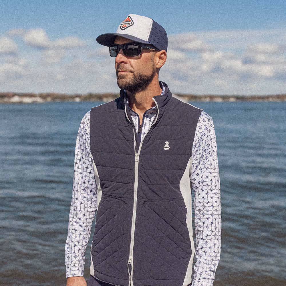 Man standing in a marina in Maryland wearing the Six Knots Every Day Performance Vest. This lightweight, navy and cream men's performance vest offers breathable comfort and versatile layering—perfect for boating, fishing, outdoor activities, and everyday coastal wear.