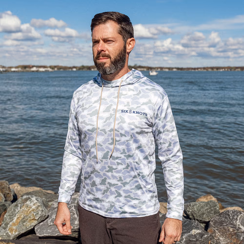 Man standing in a marina wearing the Six Knots Coastal Camo Performance Hoodie in the ‘Wetlands’ colorway — lightweight, breathable fishing hoodie with a blue and green camouflage pattern inspired by coastal wetlands; moisture-wicking and UPF sun-protective apparel ideal for saltwater fishing, boating, and all-weather marine adventures.
