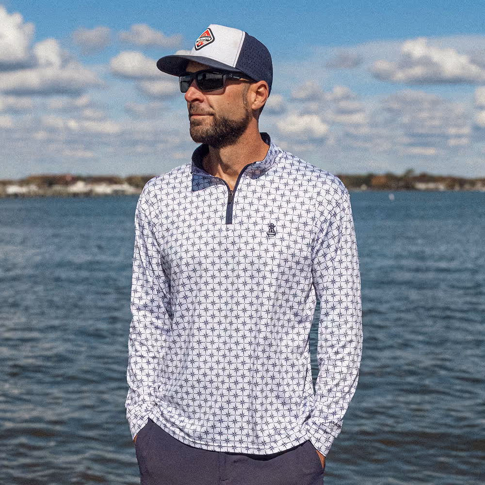 Man standing in a marina wearing the Six Knots True North Men's Performance Quarter Zip. This white quarter zip features a stylish navy compass pattern, each pointing true north — perfect for boating, coastal adventures, and sun protection in warm weather