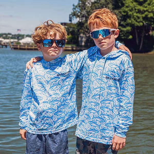 Two kids embracing while wearing Tuna Time Youth Performance Hoodies in front of the water