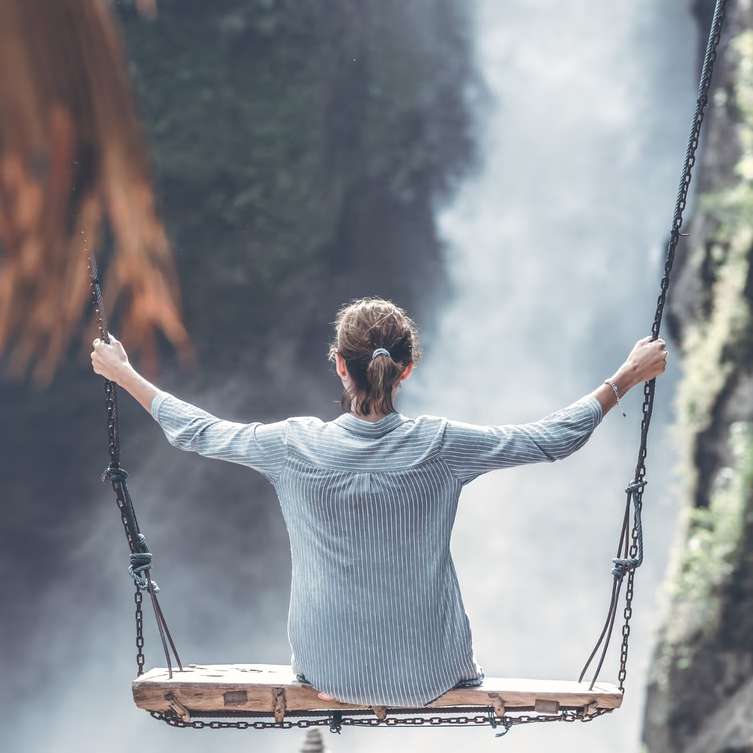 Woman sitting on a wooden swing facing a misty waterfall, holding onto the ropes with both hands, representing freedom, serenity, and courage in the face of nature.