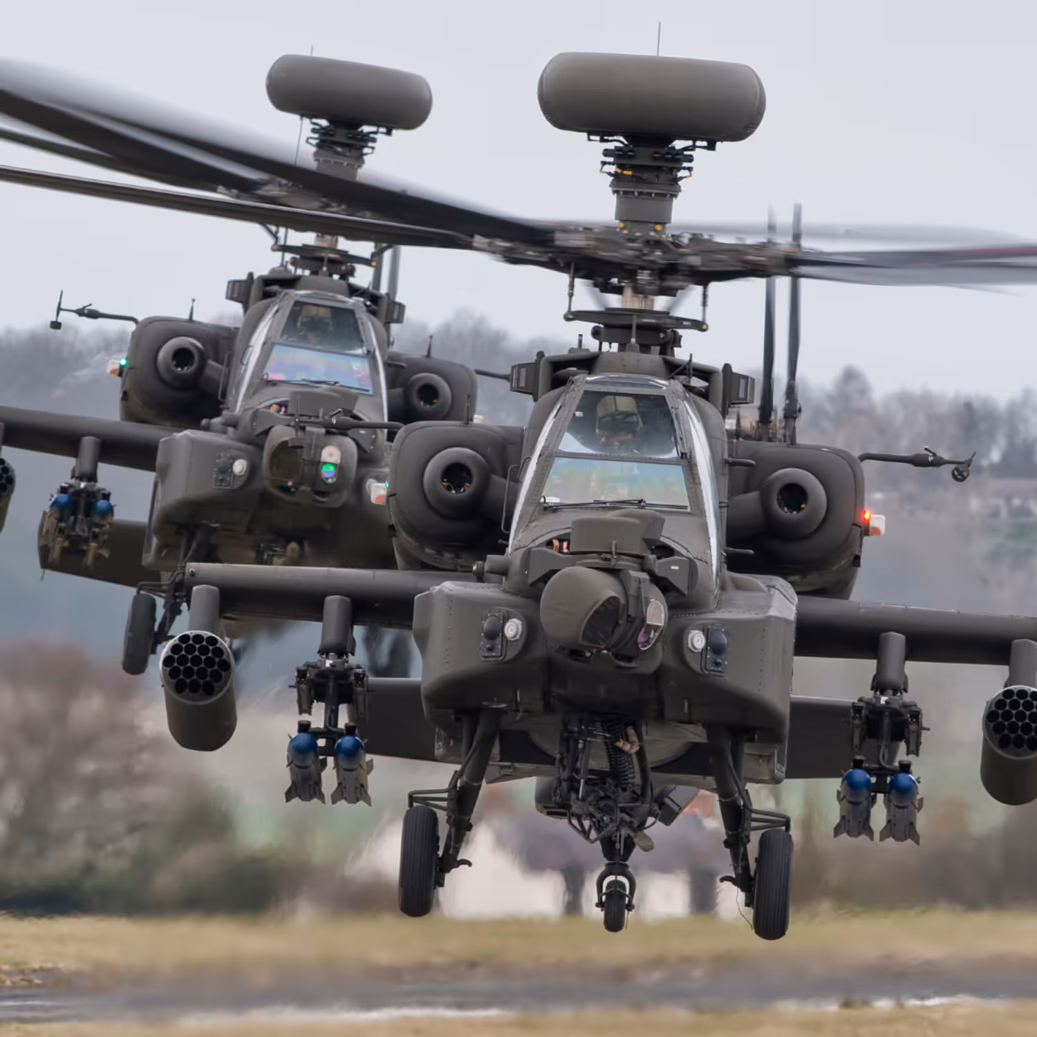 Two military AH-64 Apache helicopters in close formation, equipped with sensors, missiles, and radars, flying at low altitude over blurred terrain.