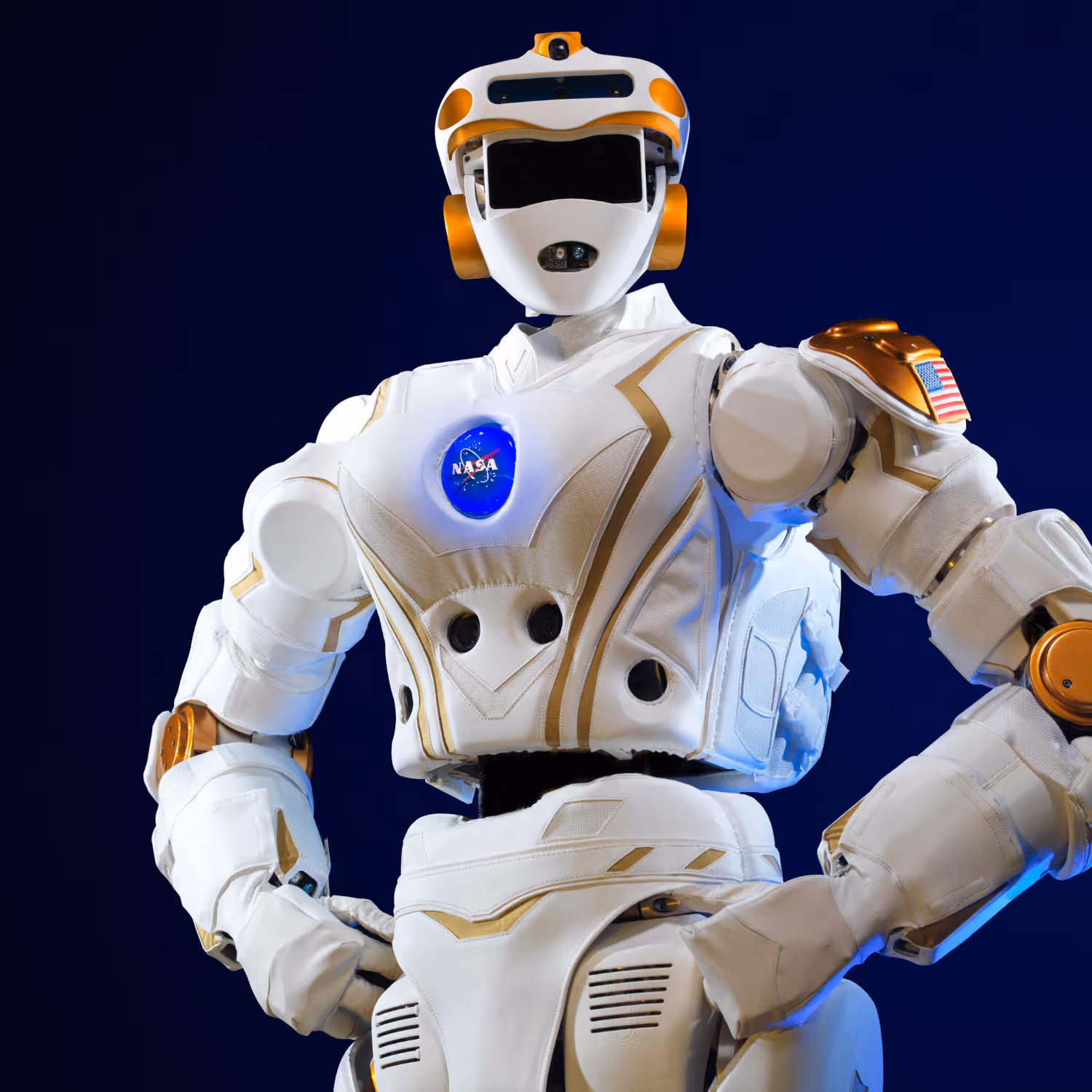 NASA’s humanoid robot "Valkyrie" standing against a dark background, featuring white and gold armor plating, the NASA logo illuminated on its chest, and an American flag patch on its shoulder.