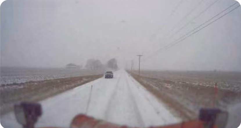 Dash cam view from a snowplow on a snow-covered rural road in falling snow