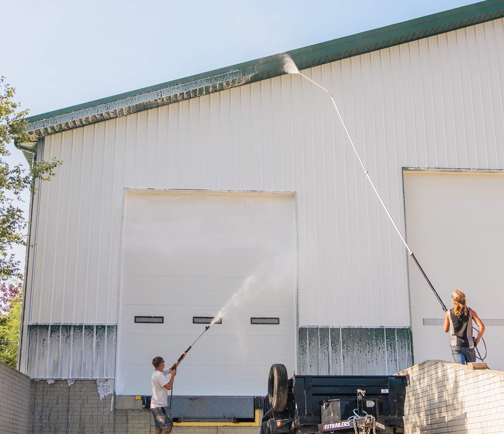 Two people power washing a large white garage door on a sunny day.