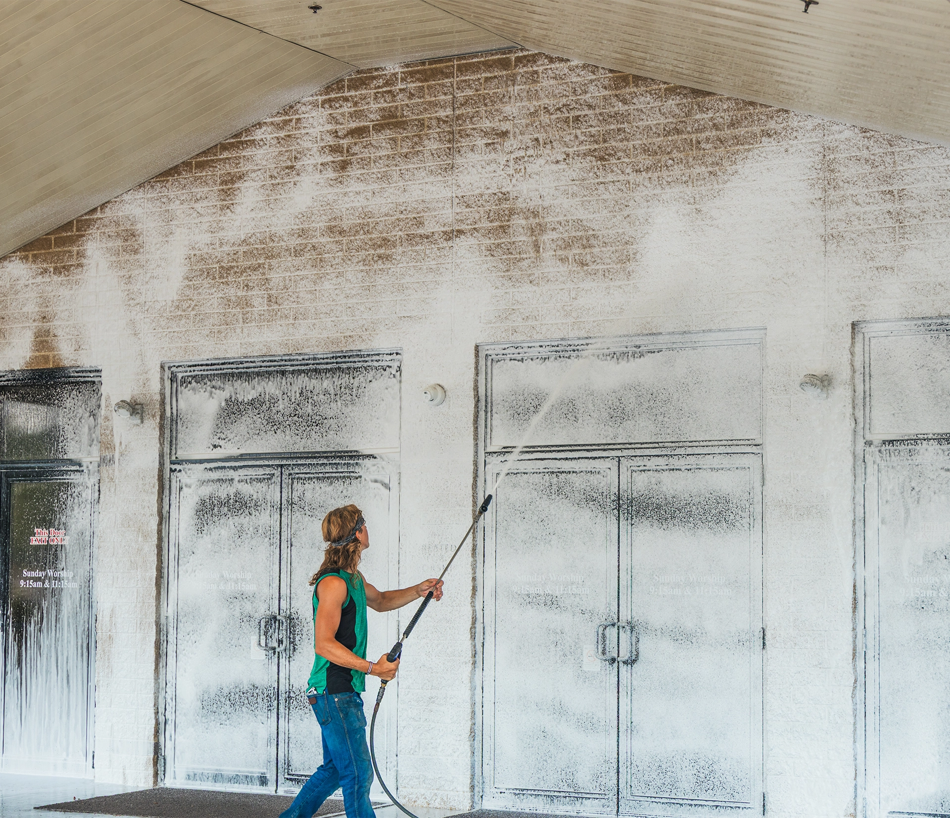Man power washing the exterior wall and doors of a large building under a beige canopy.
